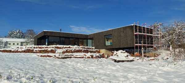 Baustellenfoto eines neugebauten Einfamilienwohnhauses mit schwarzer Holzfassade, welches im vorderen Bereich von Stahlstützen getragen ins Tal schwebt