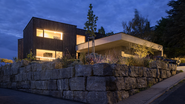 Außenaufnahme eins Architektenhauses mit schwarzer Holzfassade und Sichtbeton für die breite Doppelgarage. Der Neubau des Wohnhauses befindet sich in Schöllkrippen nahe Aschaffenburg und setzt auch architektonisch Akzente im Spessart, die über Veröffentlichungen im Cube Magazin sowie Ecotektur Magazin großen Anklang finden