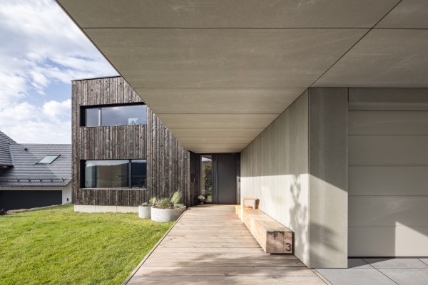 Architekturfoto vom Eingangsbereich eines neu gebauten Einfamilienhauses bei Aschaffenburg. Unter dem Betonvordach führt ein Holzsteg zum Eingang des Traumhauses, seitlich begleitet werden diese durch eine großzügige Doppelgarage in Beton