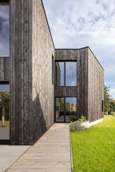 Außenaufnahme eines Wohnhaus Neubaus in Schöllkrippen nahe Aschaffenburg mit schwarzer Holzfassade, großen Glasfassaden und einem Holzsteg der von der Küche zur Terrasse führt